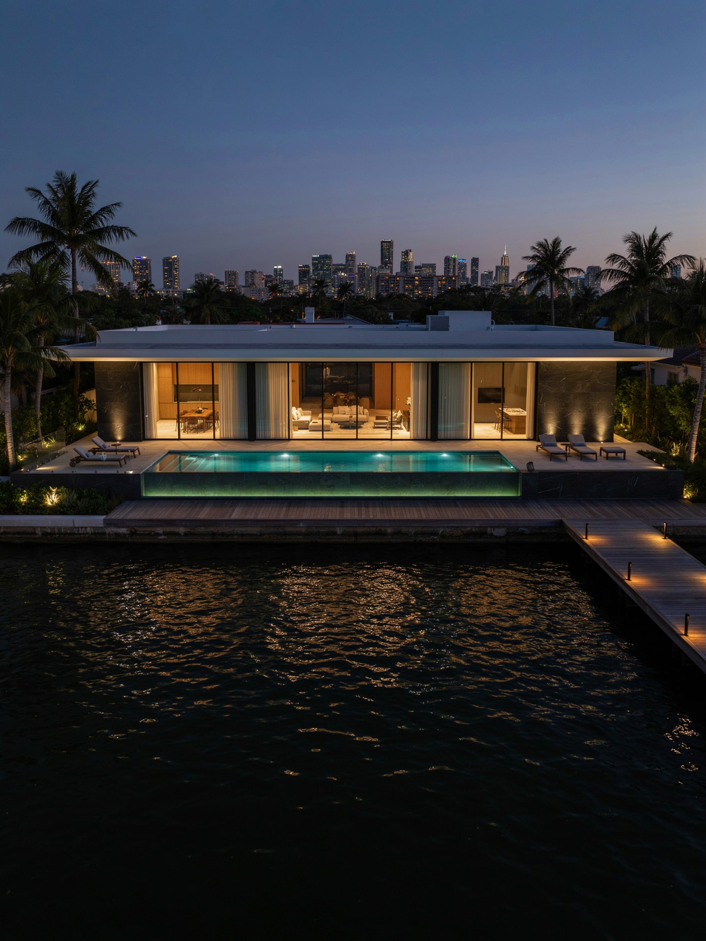 Waterfront villa at dusk with glass walls, stone cladding and a lit infinity pool